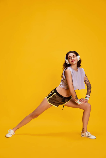 Portrait of young beautiful girl in headphones, training, doing stretching exercises isolated over yellow background. Concept of youth, beauty, sport lifestyle, emotions, facial expression. Ad - Foto, Bild
