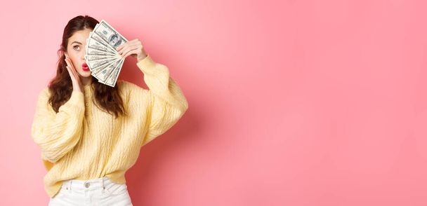 Ελκυστικό γυναικείο μοντέλο που δείχνει χρήματα, κρατώντας χαρτονομίσματα στο μισό πρόσωπο, αγγίζοντας μάγουλο και κάνοντας χείλη που φιλιούνται, στέκεται πάνω σε ροζ φόντο. - Φωτογραφία, εικόνα