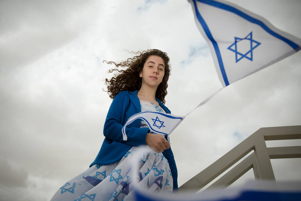 Красивая еврейская темноволосая девушка или женщина с еврейскими символами Израиля, одетые в цвета израильского флага против облачного неба снаружи, концепция Дня независимости - Фото, изображение
