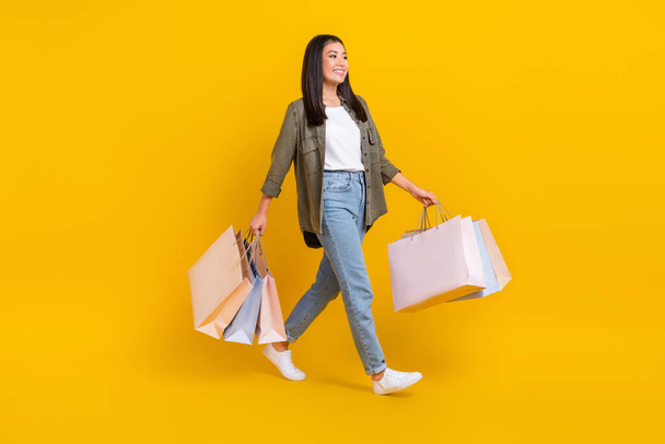 Full body photo of young positive good mood clothes designer korean girl hold packages from shopping walking boutique isolated on yellow color background. - 写真・画像