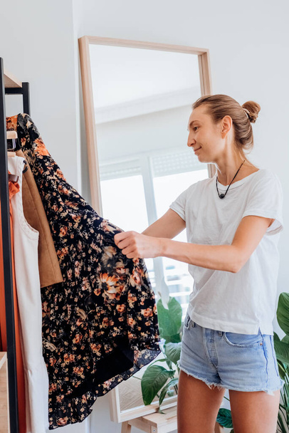Blonde young woman choosing clothes on clothes rack dressing looking herself in mirror. Girl think what to wear. Lifestyle women. High quality photo - Φωτογραφία, εικόνα