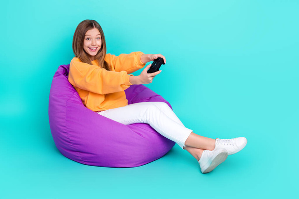 Full length photo of pretty cute school girl dressed orange hoodie bean bag having fun playstation empty space isolated teal color background. - Foto, imagen