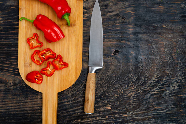 Fresh raw sweet pepper (bell pepper) on a cutting board with a knife. Chopped pepper pieces are ready to cook. Dark wooden table, top view.Space for text. - Foto, Imagen