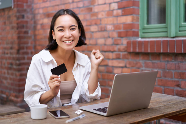 Happy young asian woman sitting near laptop, holding credit card, paying bills, shopping online contactless, smiling at camera. - Foto, Imagen