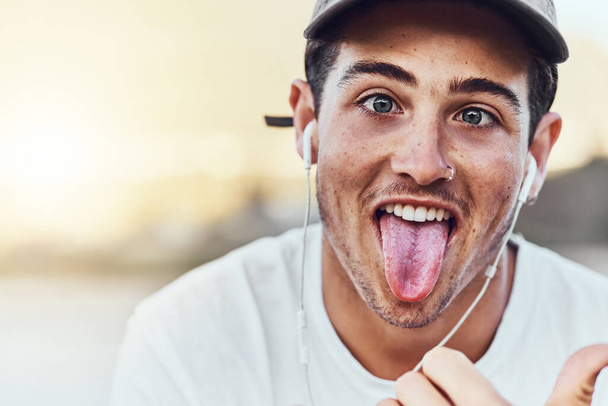Fashion, city and portrait man with tongue out enjoy weekend, summer vacation and holiday. Freedom, comic and young male with funny face expression listening to music, radio and audio in urban town. - Photo, Image