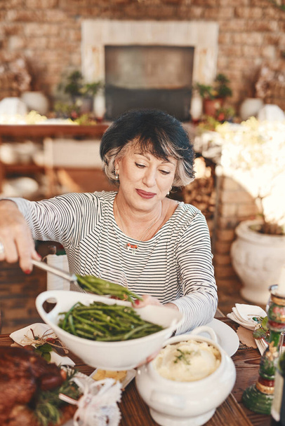 感謝祭のテーブルとアメリカのお祝いのためのフードパーティーで提供する野菜と高齢者の女性。休日、栄養のための緑豆と自宅のダイニングテーブルでお祝いと成熟したお祝いの人 - 写真・画像
