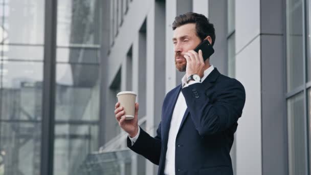 Executive businessman konsultant menedżer sprzedaży rozmawiając przez telefon komórkowy zdalnie doradzając klientowi pozytywny zajęty człowiek stojący na zewnątrz przy przerwie pijąc kawę na czacie z przyjacielem za pomocą smartfona - Materiał filmowy, wideo