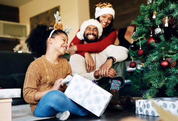 Μητέρα, πατέρας και κορίτσι με ένα χριστουγεννιάτικο δώρο αγκαλιάζει για να γιορτάσουν την αγάπη, ευτυχισμένη οικογένεια και τις χειμερινές διακοπές στο σπίτι. Με αγάπη, μαμά και ενθουσιασμένο μπαμπά με το καπέλο του Άι Βασίλη να δίνουν σε ένα μικρό παιδί ή παιδί ένα κουτί έκπληξη. - Φωτογραφία, εικόνα