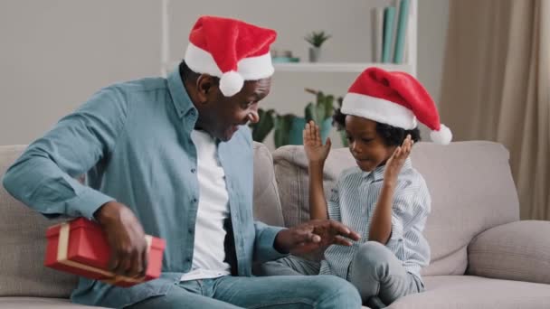 Gelukkig Afrikaans Amerikaans gezin vieren Nieuwjaar liefdevolle vader geeft dochter kerstcadeau schattig klein kind meisje vreugde in verrassing kind knuffels vader zitten in gezellige kamer op de bank genieten van vakantie - Video