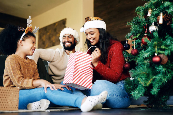 Χριστούγεννα, αγάπη και οικογένεια ανοίγοντας δώρα στο σπίτι, διασκεδάζοντας και δένοντας στις διακοπές. Χριστούγεννα πνεύμα, φροντίδα και ευτυχισμένη πατέρα, μητέρα και κορίτσι ανταλλαγή δώρα στο σαλόνι και απολαμβάνοντας ποιοτικό χρόνο - Φωτογραφία, εικόνα