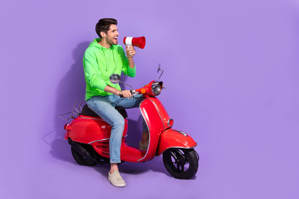 Full size photo of satisfied activist guy green hoodie hold loudspeaker look empty space on motorcycle isolated on violet color background. - Fotoğraf, Görsel