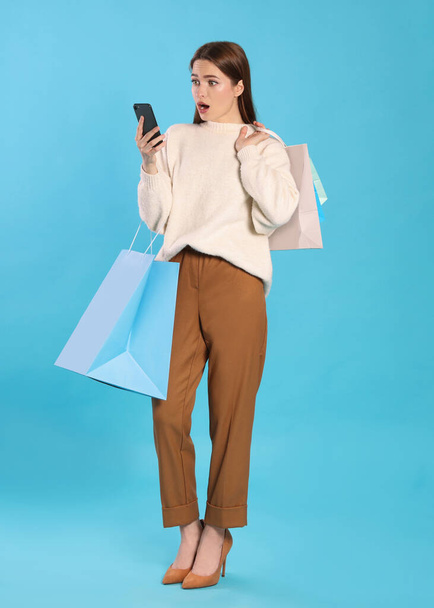 Surprised young woman with shopping bags looking at smartphone on light blue background. Big sale - Foto, immagini
