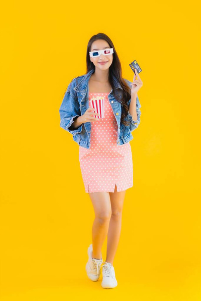 Портрет красивая молодая азиатская женщина готова смотреть кино с попкорном и кредитной карты на желтом изолированном фоне
 - Фото, изображение