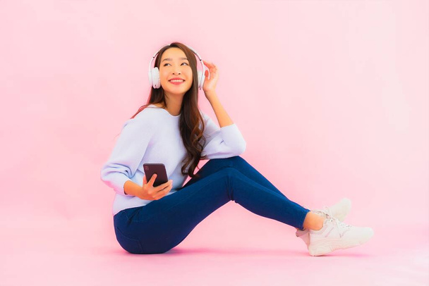 Retrato hermosa joven asiática mujer uso inteligente teléfono móvil con auriculares para escuchar música sobre fondo rosa - Foto, imagen