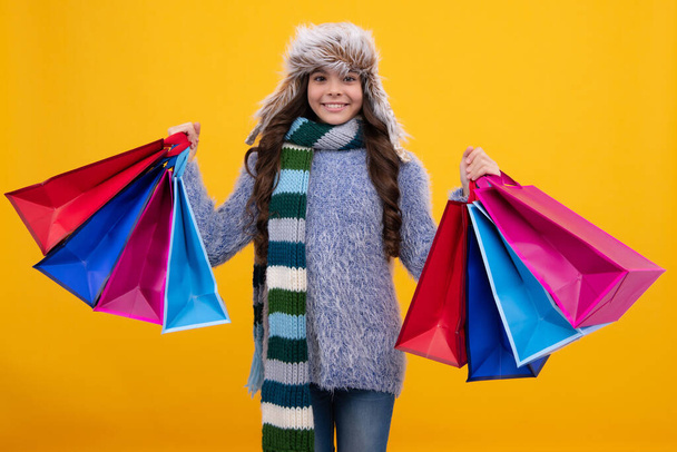 Teenager child girl in winter hat with shopping bags on yellow background. Kid with shopping winter sale bags - Fotografie, Obrázek