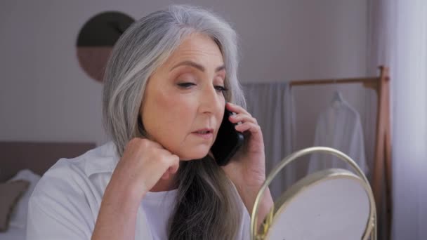 Hablar de teléfono hablar móvil hablar senior gris de pelo viejo 60s mujer señora mirando espejo reflexión tocar la cara arrugado piel hablar remoto conversación envejecimiento perfecto suave cuidado de la piel hidratante - Imágenes, Vídeo