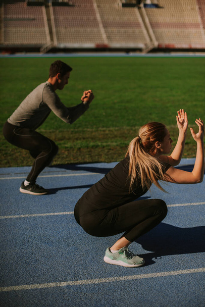 Attractive couple is working out together. They are doing squats, on a sunny morning at the stadium - Photo, Image