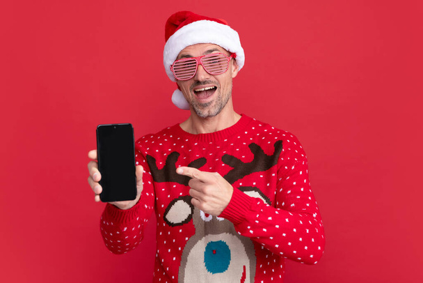 překvapený muž ve svetru a klobouku Santa Clause. Vánoční chlap ukazuje prstem na obrazovku telefonu na červeném pozadí. Šťastný nový rok. Veselé Vánoce. pozdravy online. - Fotografie, Obrázek
