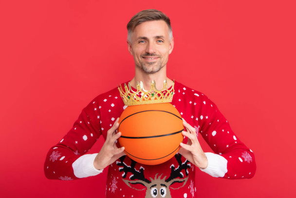 Wintermann mit Krone hält Basketballball. Mann in Weihnachtspullover und Hut auf farbigem Hintergrund. Weihnachtsmann mittleren Alters - Foto, Bild