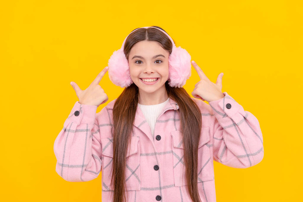 Šťastná dívka dítě úsměv ukazuje prsty na teplé zimní kožešiny ohřívače uší žluté pozadí, chrániče uší. - Fotografie, Obrázek