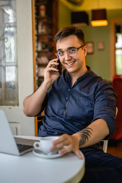 Jeden mężczyzna młody biały mężczyzna siedzi w biurze w domu lub w kawiarni pracując na laptopie sam uśmiech freelance projekt prawdziwi ludzie kopiować przestrzeń za pomocą telefonu komórkowego rozmowa wykonując połączenie - Zdjęcie, obraz