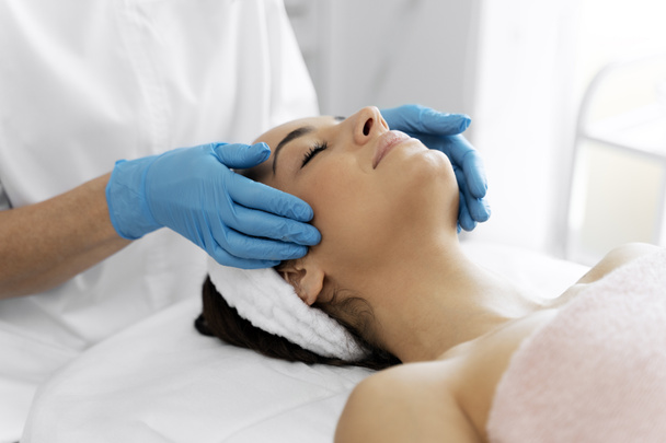 Agradable procedimiento de belleza con suave masaje facial realizado por el terapeuta profesional. Mujer joven tendida con los ojos cerrados  - Foto, imagen