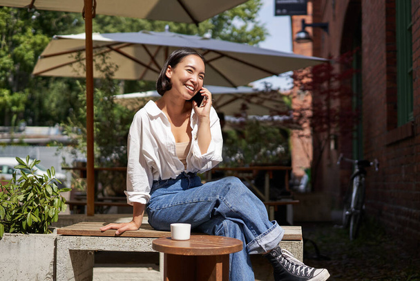 Люди і спілкування. Стильна азіатська дівчина сидить у відкритому кафе з чашкою кави та смартфона, використовуючи мобільний телефон
. - Фото, зображення