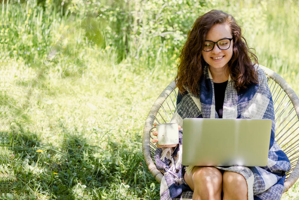 Ευτυχισμένος νεαρός ενήλικας σγουρά μαλλιά γυναίκα φορώντας γυαλιά χρησιμοποιεί ένα φορητό υπολογιστή, ενώ κάθεται σε ένα καταπράσινο κήπο στην ύπαιθρο. - Φωτογραφία, εικόνα