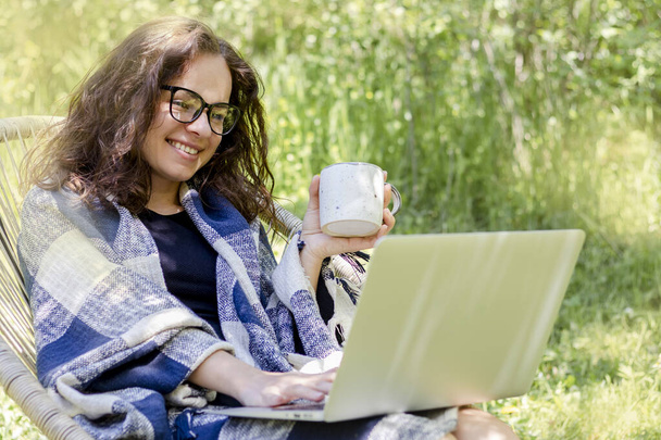 Nő kockás csomagolásban használja a laptopját és kávét iszik a kertben. - Fotó, kép