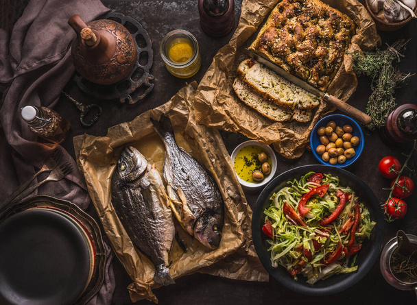 Mediterranean lunch or dinner with roasted dorado fishes, homemade focaccia bread , olive oil and olives served on rustic table with tableware and kitchen utensils, top view. - Foto, Bild