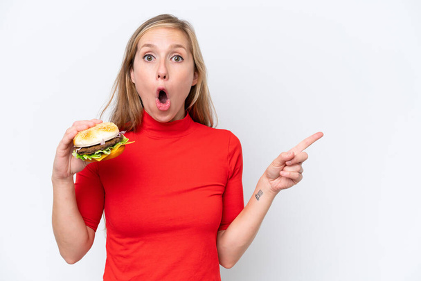 Młoda blondynka trzyma hamburgera odizolowany na białym tle zaskoczony i wskazując bok - Zdjęcie, obraz