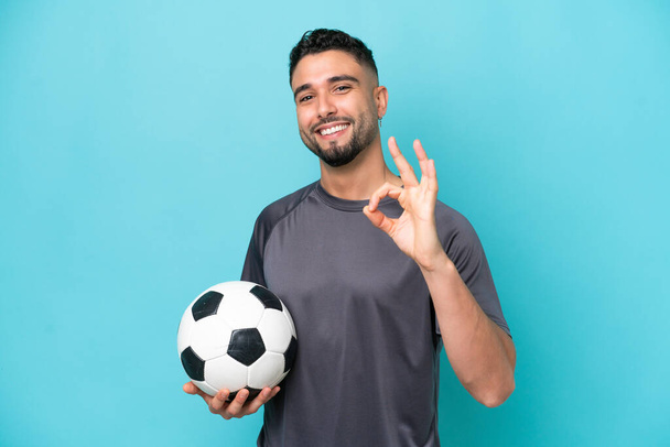 Young Arab handsome man isolated on blue background with soccer ball and making OK sign - Photo, Image