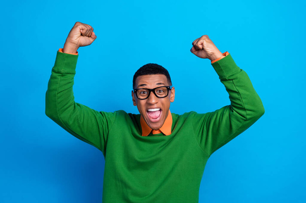 Photo of optimistic satisfied man wear orange shirt under green sweatshirt raise fists up scream yell isolated on blue color background. - Photo, Image