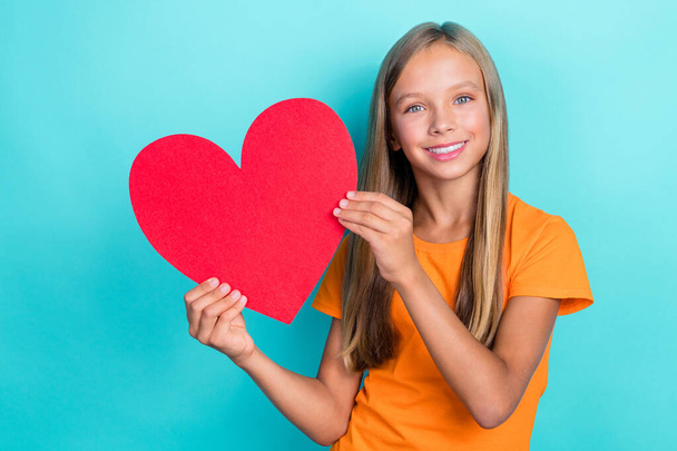 Closeup portrait photo of preteen schoolkid girl wear orange t-shirt blonde hair hold red paper heart toothy smile good mood isolated on cyan color background. - Foto, imagen