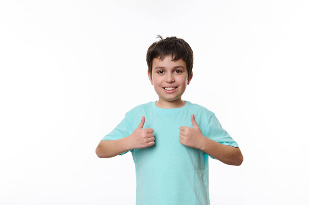 Retrato de branco bonito menino adolescente agradável em azul casual t-shirt, sorrindo agradavelmente, olhando para a câmera, mostrando polegares para cima, isolado em fundo branco com espaço para texto promocional - Foto, Imagem