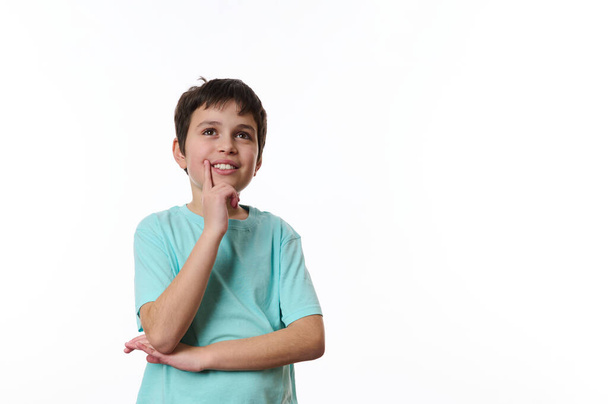 Gelukkige tiener jongen draagt turquoise t-shirt, houdt zijn hand op kin en schattig glimlachen, bedachtzaam kijken naar opzij, geïsoleerd over witte achtergrond met kopieeradvertentie ruimte voor promotietekst - Foto, afbeelding