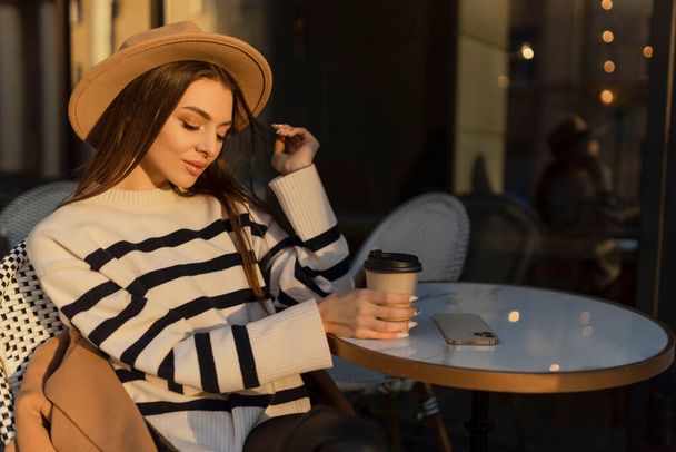 beautiful smiling woman in stylish outfit sitting at the table in a hat and sweater, romantic happy mood, waiting for a boyfriend on a date in a cafe, spring-summer fashion trend, drinking coffee. - Photo, Image