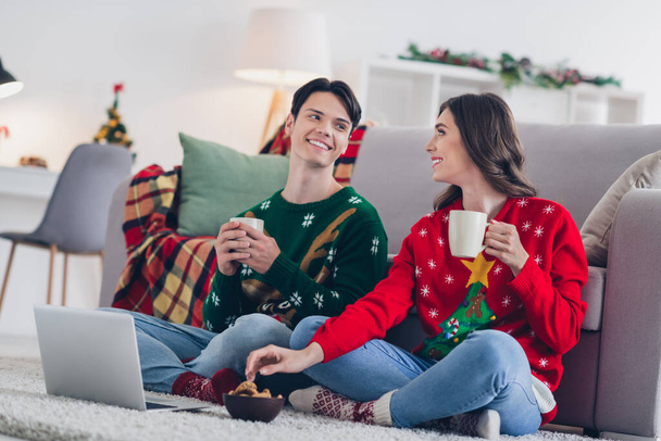 Full length photo of two peaceful idyllic people sit carpet floor hold hot chocolate tea mug eat sweet cookies communicate watch film indoors. - 写真・画像