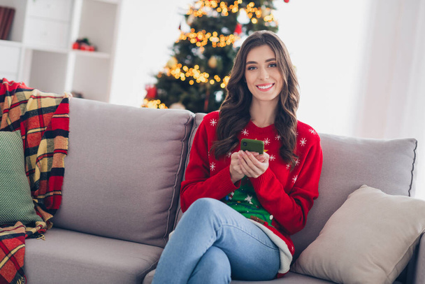 Photo of cute lovely smiling positive lady sitting relaxed favorite rest room sofa enjoy christmas time with smartphone new xmas tree indoors. - Фото, изображение