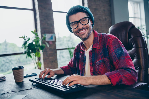 Photo of happy smiling coder dressed hat glasses typing program modern gadget indoors workplace workstation loft. - Foto, imagen