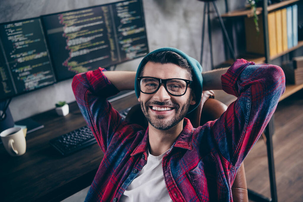 Photo of good mood happy freelancer wear hat glasses hands arms behind head smiling indoors workplace workstation loft. - Foto, Imagem