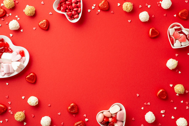 Valentine's Day concept. Top view photo of heart shaped saucers with confectionery candies and confetti on isolated red background with blank space in the middle - Photo, image