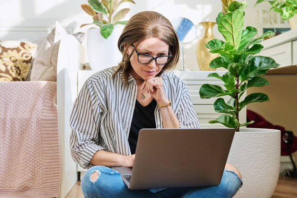 Mature woman sitting on the floor at home looking at a laptop. Middle age female using laptop for business work leisure shopping communication entertainment - Foto, Bild