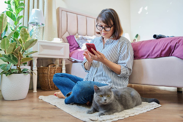 Home lifestyle, woman with cat, comfort calmness concept. Female sitting on floor on carpet using smartphone, pet gray british cat lying near owner - Zdjęcie, obraz