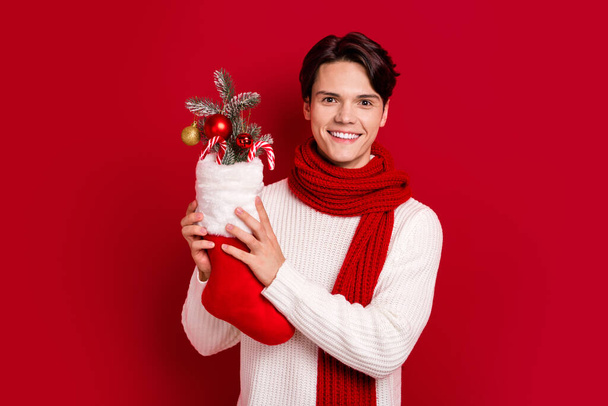 Photo of young smile brunet hair student guy wear fall season scarf hold xmas sock adornment baubles preparation isolated on red color background. - Fotó, kép