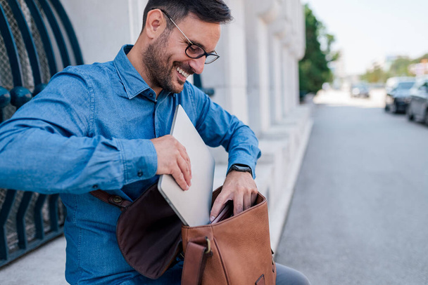 Hombre adulto alegre, terminado con su trabajo, guardando su portátil, sentado afuera en la ciudad. - Foto, Imagen