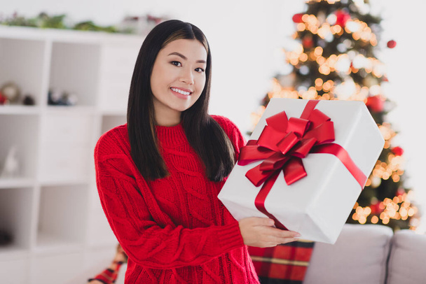 Portrait of good mood gorgeous toothy beaming woman with straight hairdo wear red knit pullover holding present box in house indoors. - Foto, Imagen