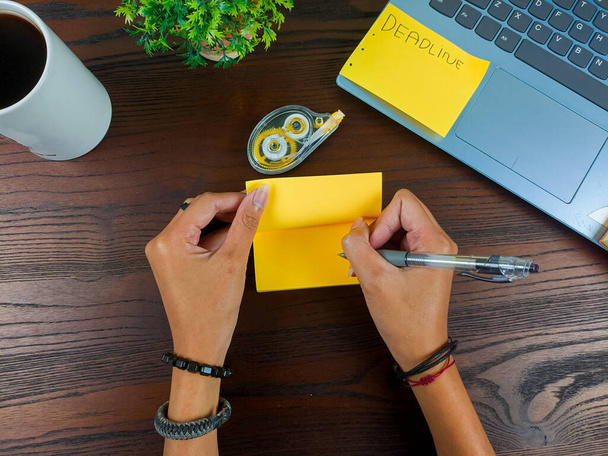 le donne scrivono in note gialle, fingono note gialle. Le mani delle donne scrivono appunti su carta da lettere gialla sullo sfondo dell'area di lavoro della scrivania dell'ufficio dalla vista dall'alto. - Foto, immagini