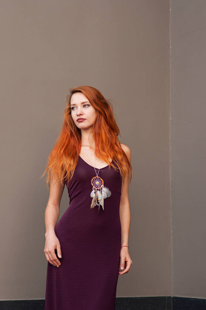 Portrait of beautiful slim redhead girl in wine boho chic dress and handmade white dreamcatcher feather talisman pendant - Photo, Image