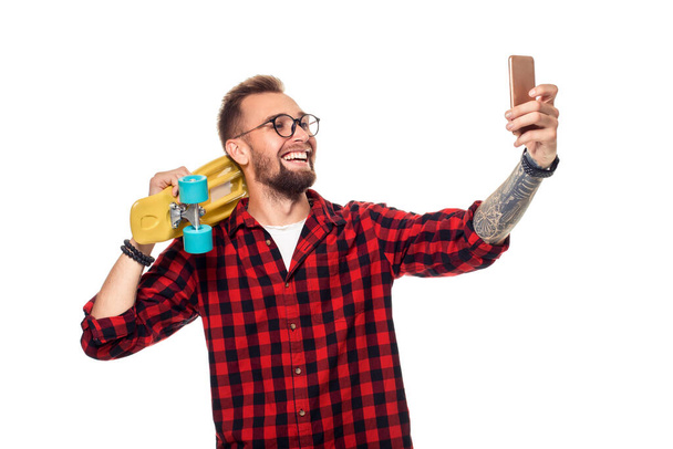 Young man holding the skateboard on shoulder raising his phone takes a selfie on a white background. Carefree youth and freedom. Active guy in plaid shirt with copy space - Foto, afbeelding